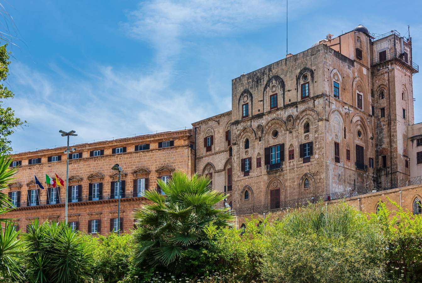 Extérieur du palais aux murs beiges, fenêtres cintrées et tour sous un ciel bleu.