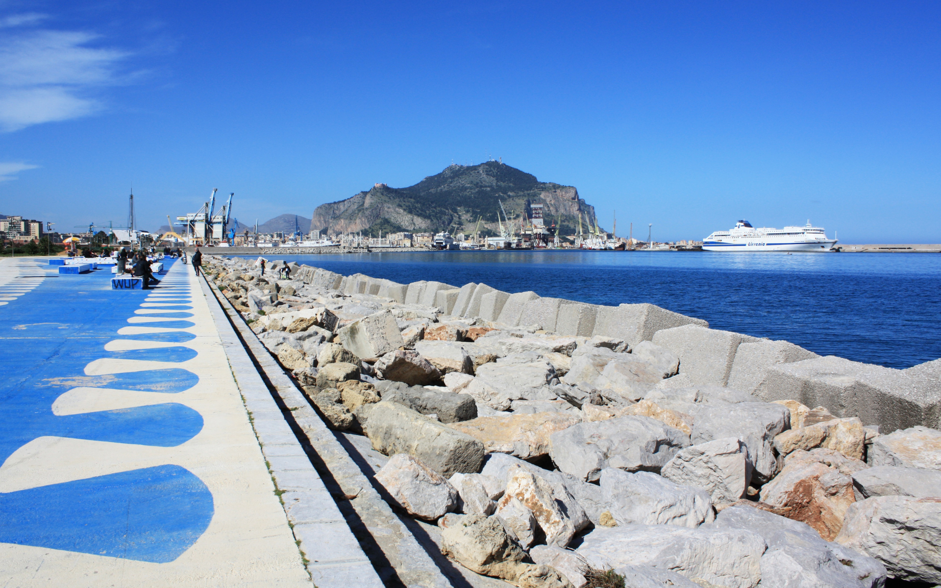 Promenade en bord de mer avec des touches de bleu et de blanc, une montagne et un ferry par une journée ensoleillée.