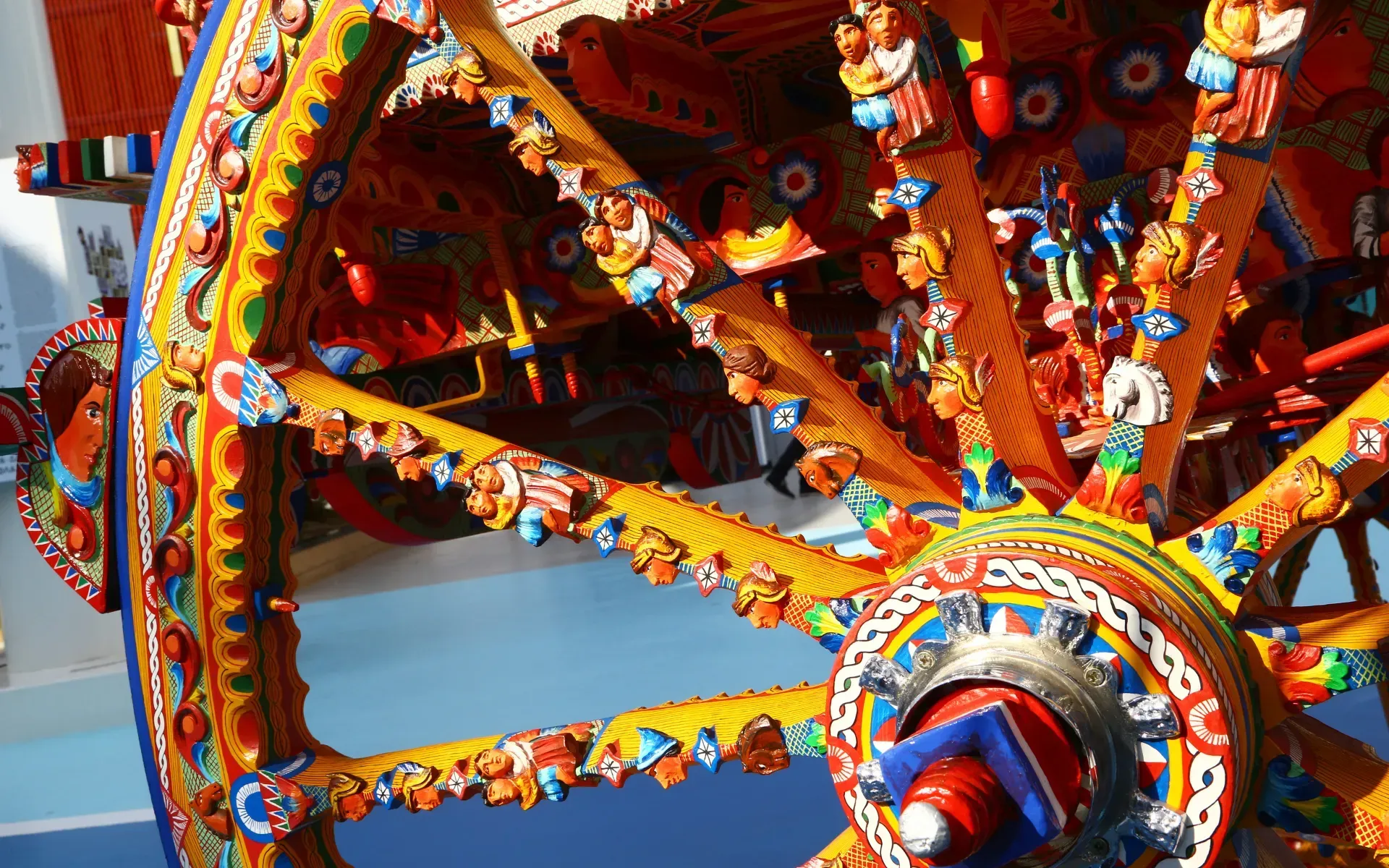 Gros plan sur une roue de charrette sicilienne aux couleurs vives, ornée de sculptures complexes jaunes, oranges, bleues et rouges.