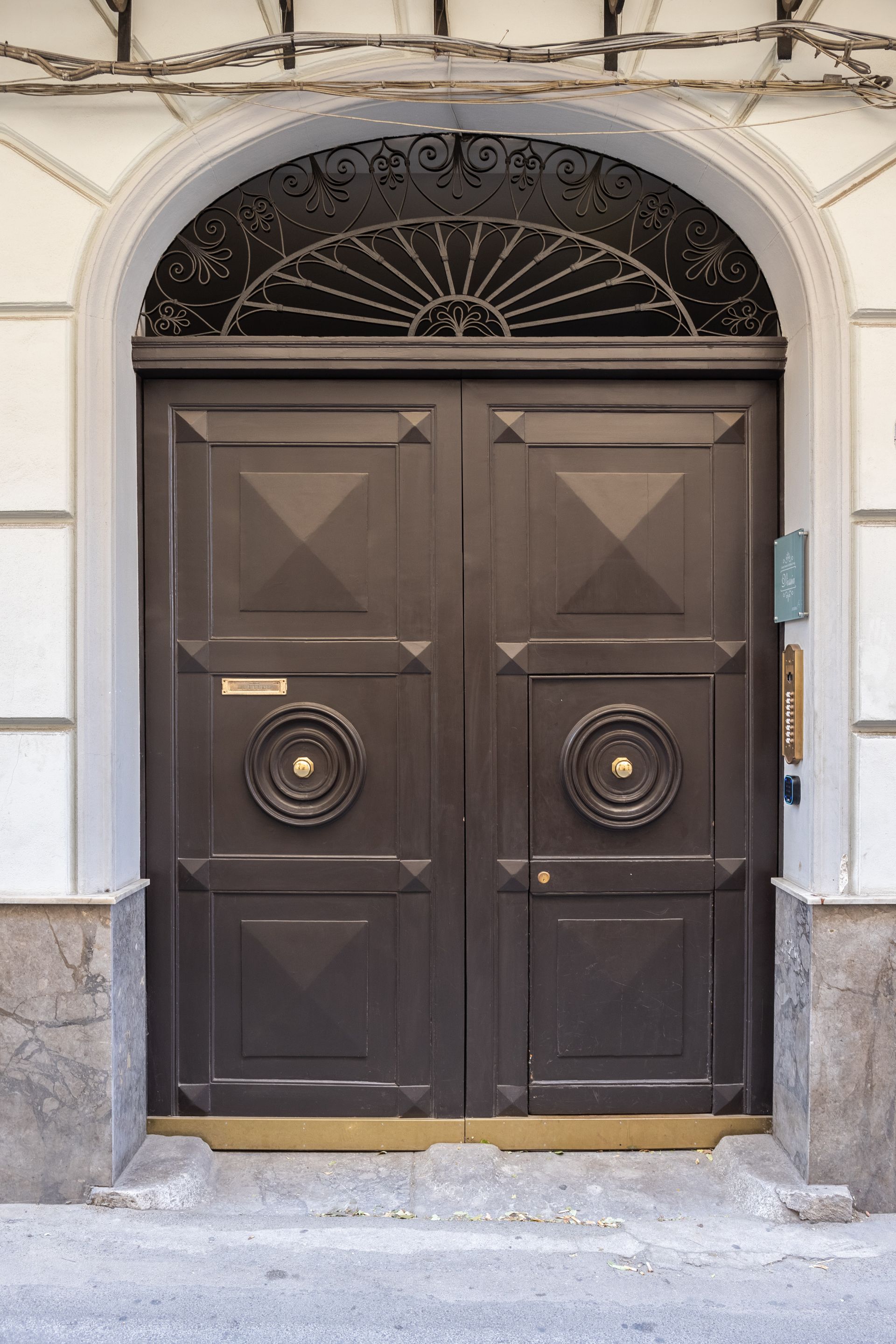Doppie porte marrone scuro con ferramenta dorata e parte superiore arcuata, sotto una decorazione in metallo.