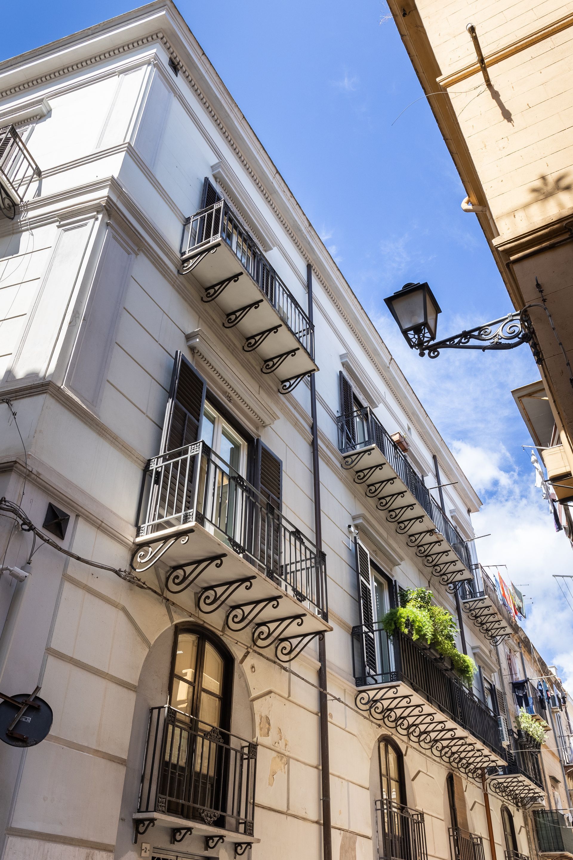 Edificio bianco con balconi in ferro battuto decorati; il cielo azzurro fa capolino dalla stretta strada.