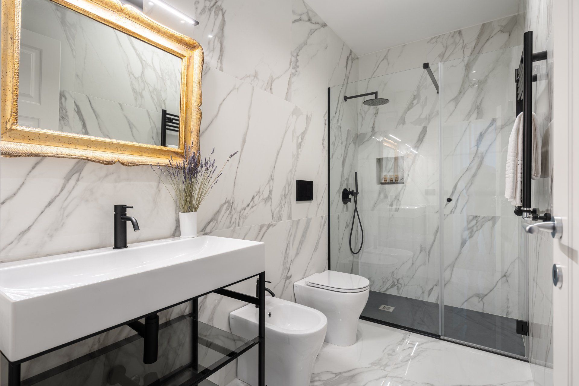 Bagno moderno con pareti in marmo bianco, specchio con cornice dorata, sanitari neri, doccia in vetro e mobile lavabo bianco.