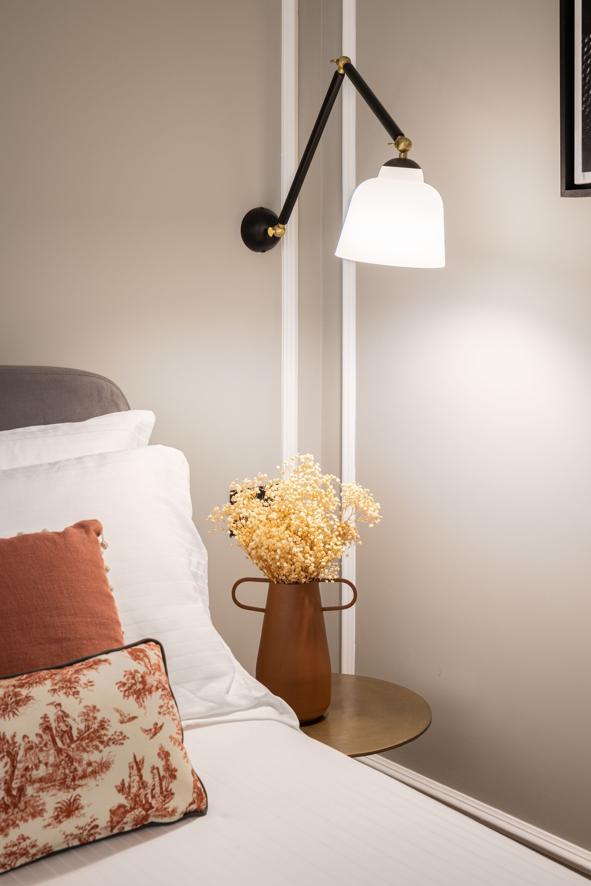 Angolo della camera da letto con lampada a parete, vaso di fiori secchi sul comodino e cuscini sul letto.