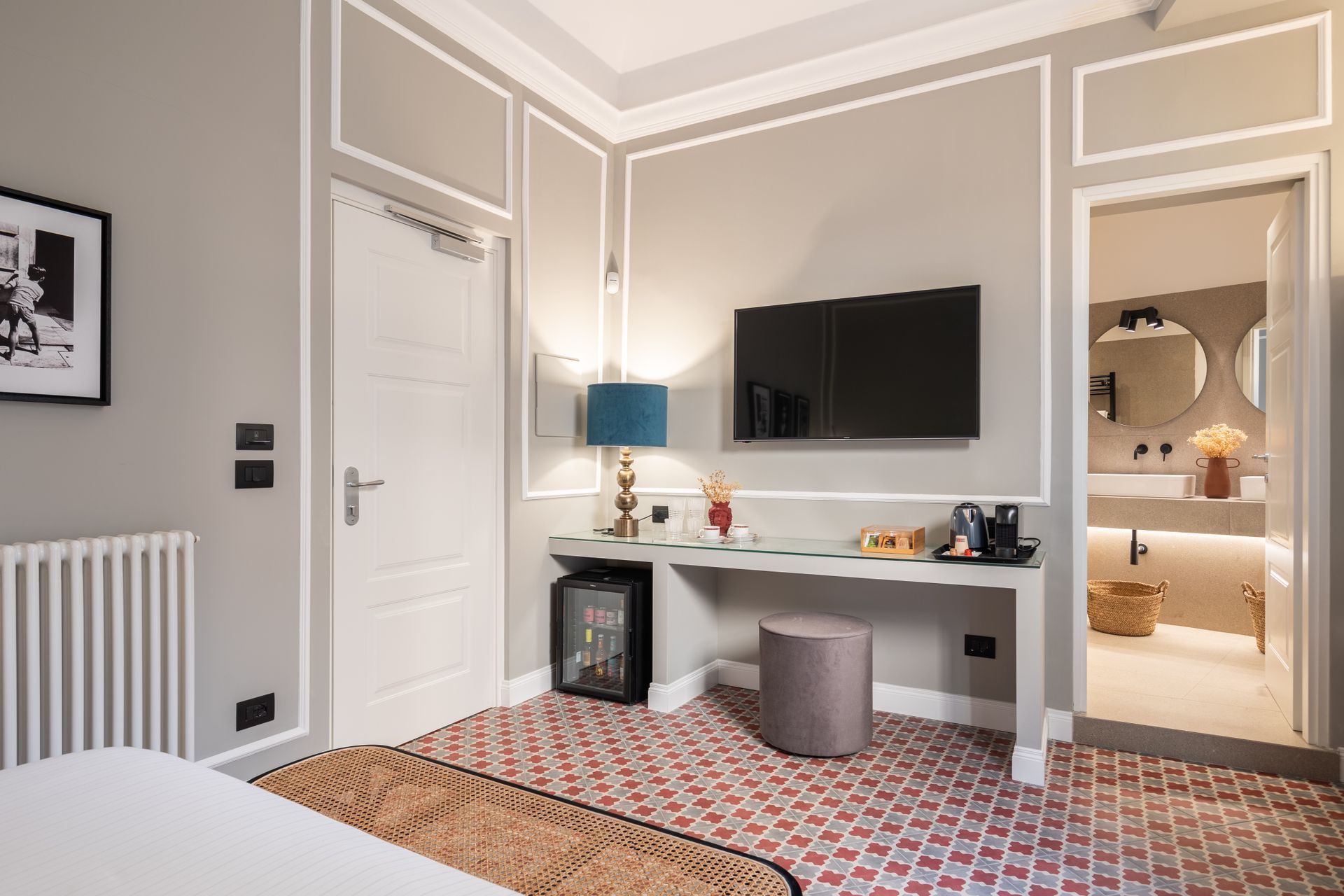 Camera d'albergo con pareti grigie, scrivania, TV e bagno a vista. Pavimento in piastrelle rosse.
