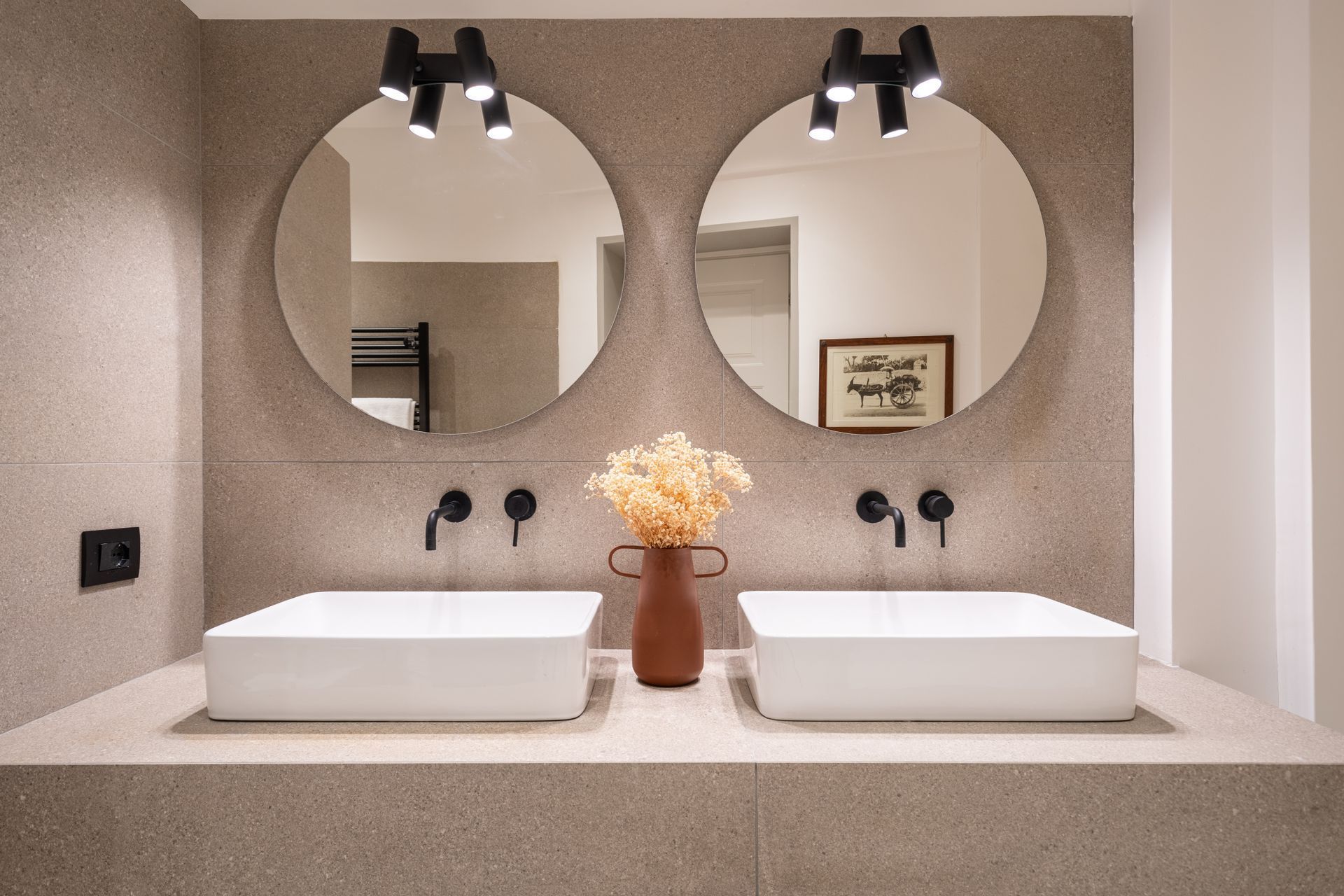 Bagno con due lavandini bianchi, specchi rotondi e sanitari neri contro una parete beige testurizzata.