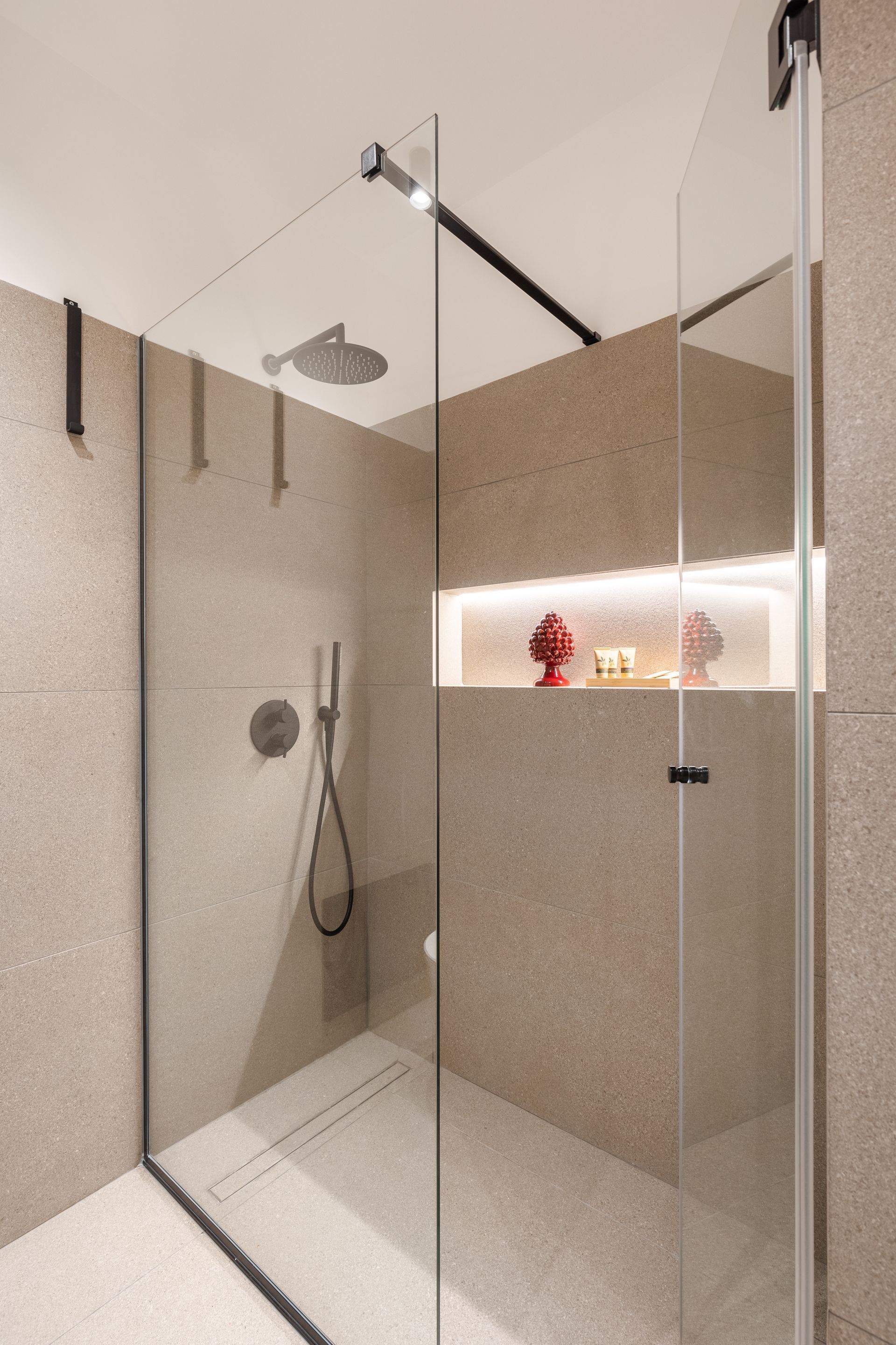 Doccia walk-in con porte in vetro, pareti in pietra, soffione a pioggia e mensola incassata con statuette rosse.