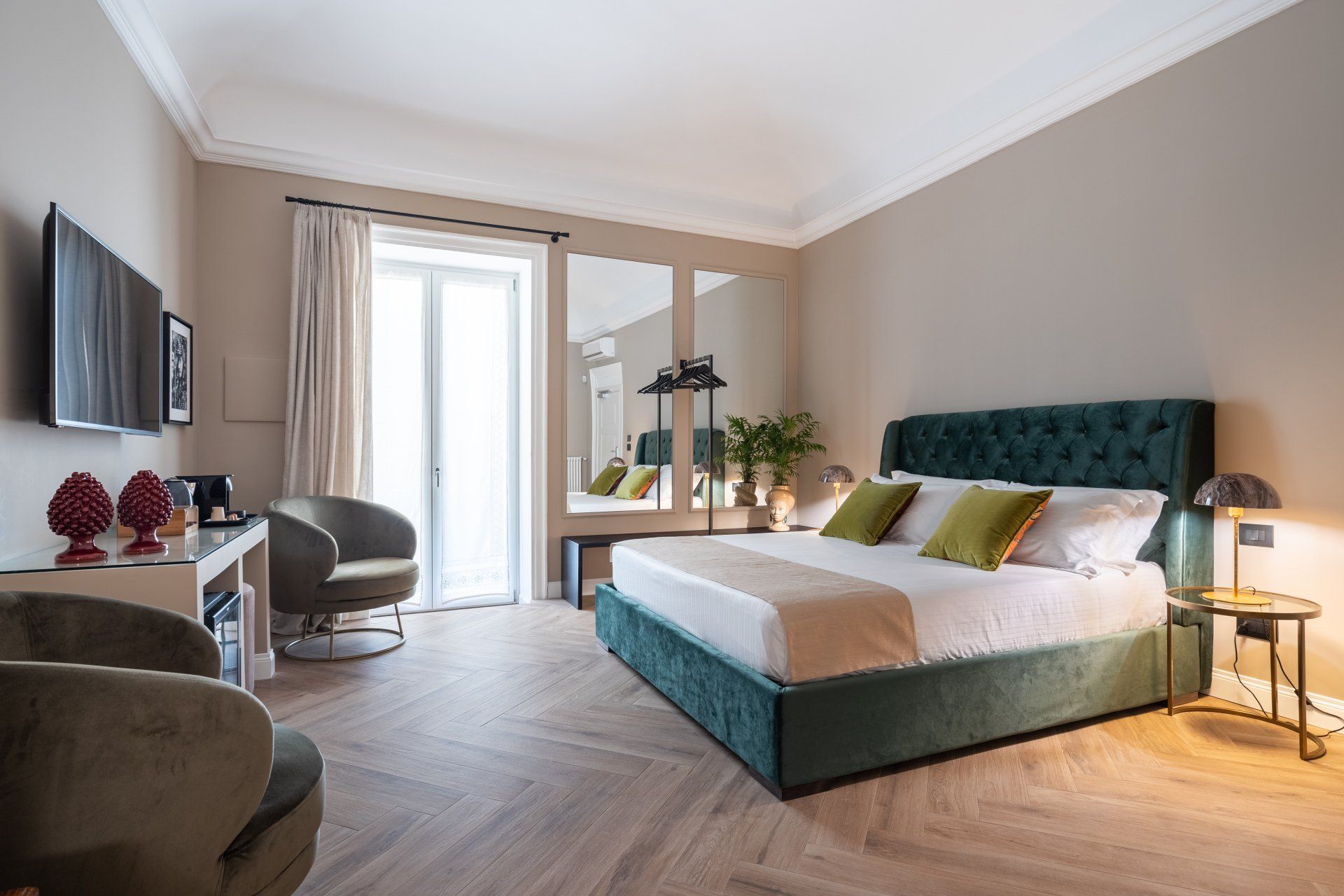 Chambre avec un lit capitonné vert, deux fauteuils, une télévision et un grand miroir. Murs beiges et parquet.