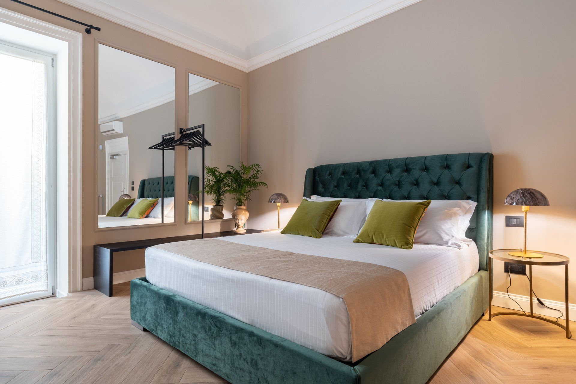 Un lit vert dans une chambre aux tons neutres, flanqué de miroirs, de plantes et d'une table de chevet avec une lampe.