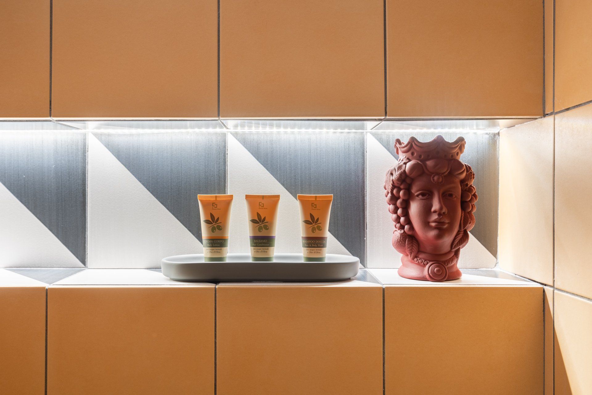 Mensola con articoli da toeletta, un busto e uno sfondo di piastrelle geometriche. Colori crema, arancione e nero.