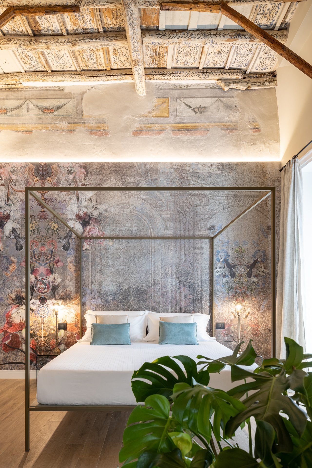 Chambre avec lit à baldaquin, papier peint fleuri et plafond à poutres apparentes. Coussins turquoise et plante d'intérieur au premier plan.