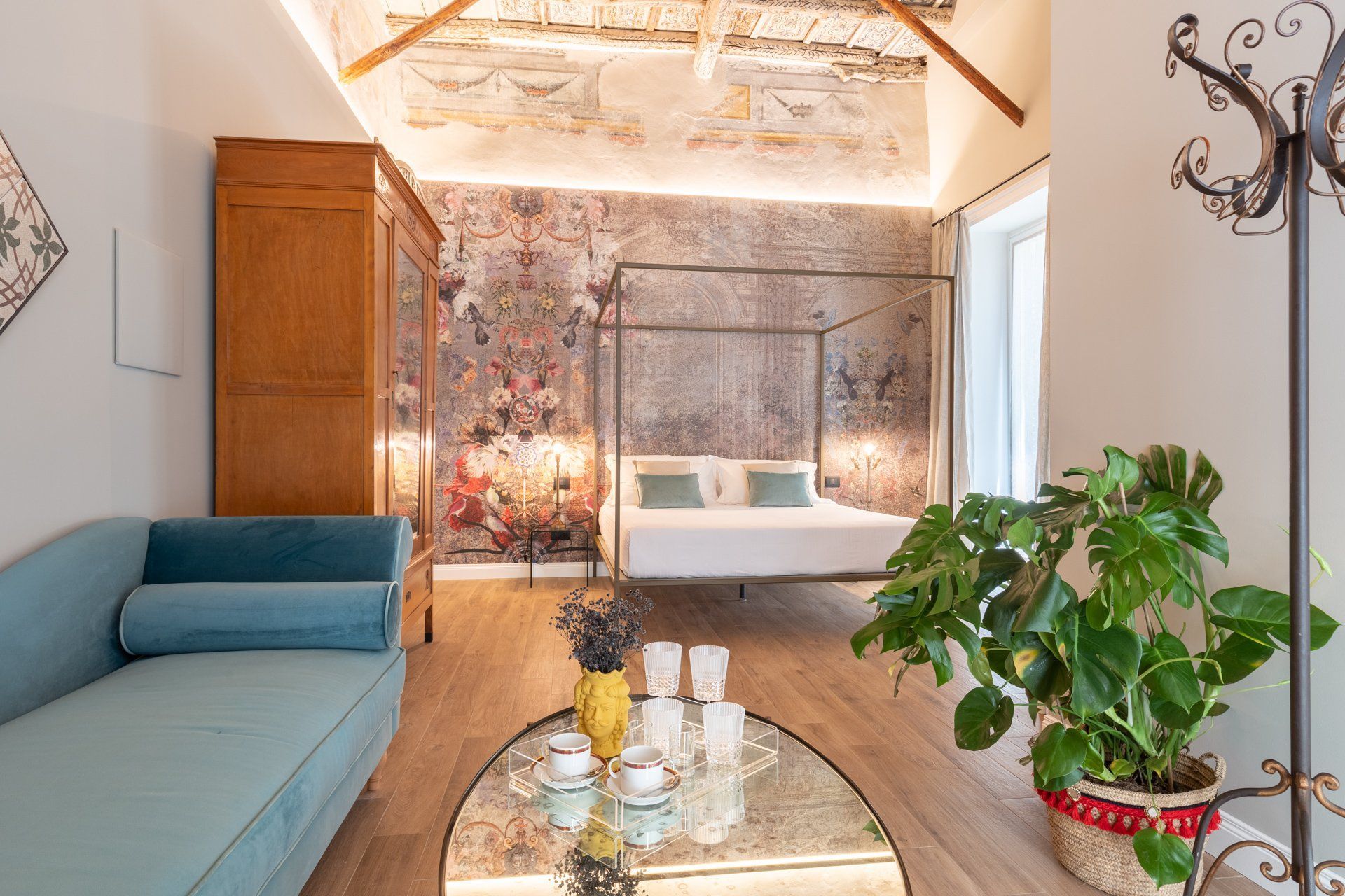 Soggiorno con divano blu, armadio in legno, letto a baldacchino, carta da parati floreale e grande pianta.