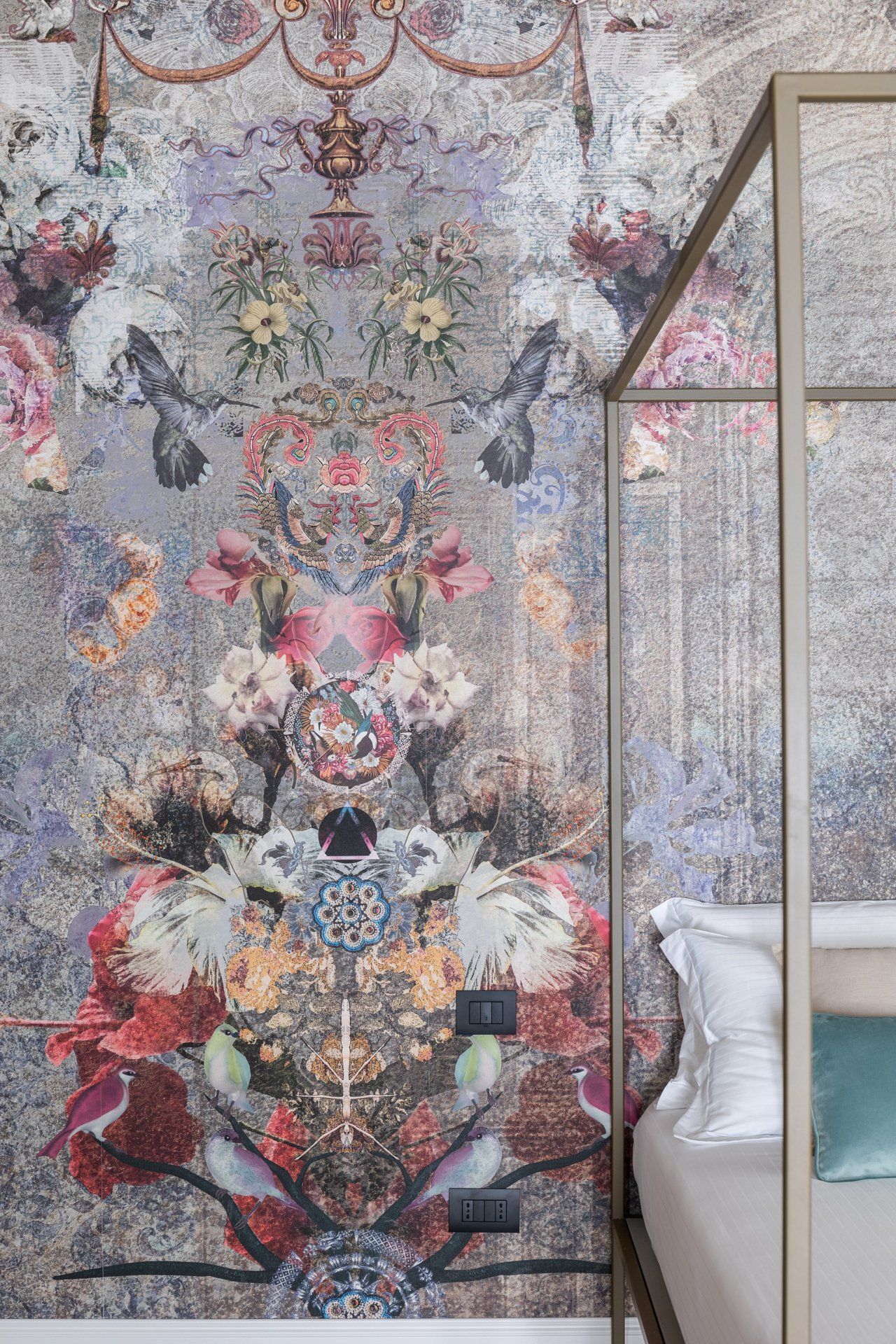 Una carta da parati floreale con uccelli decora una parete accanto a un letto beige con cornice dorata e cuscini bianchi.