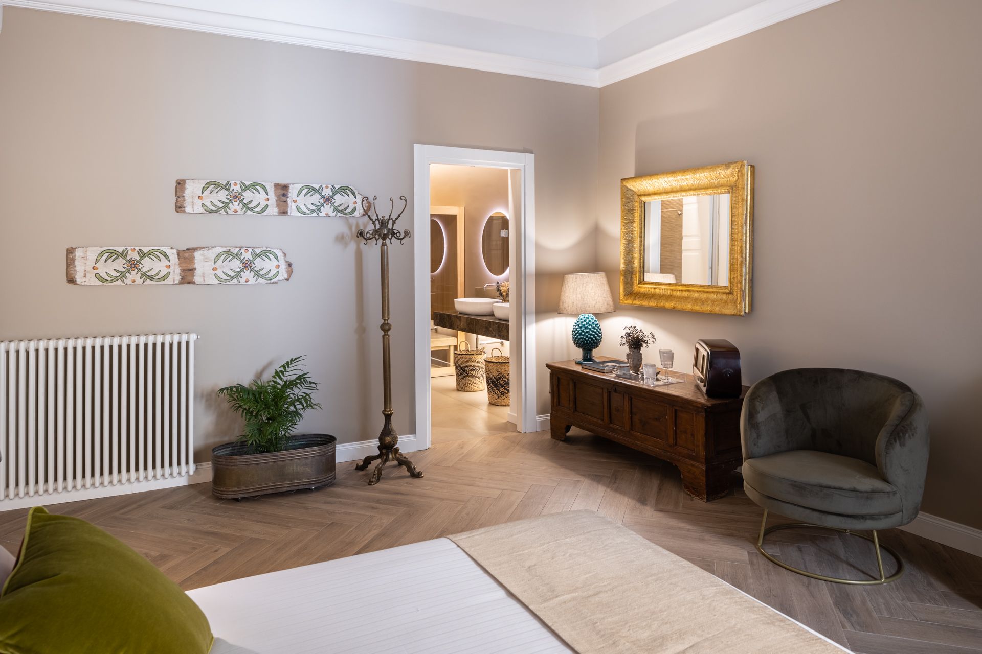 Bedroom with tan walls, herringbone wood floor, and doorway to a bathroom. Includes a radiator, artwork, chair, and dresser.