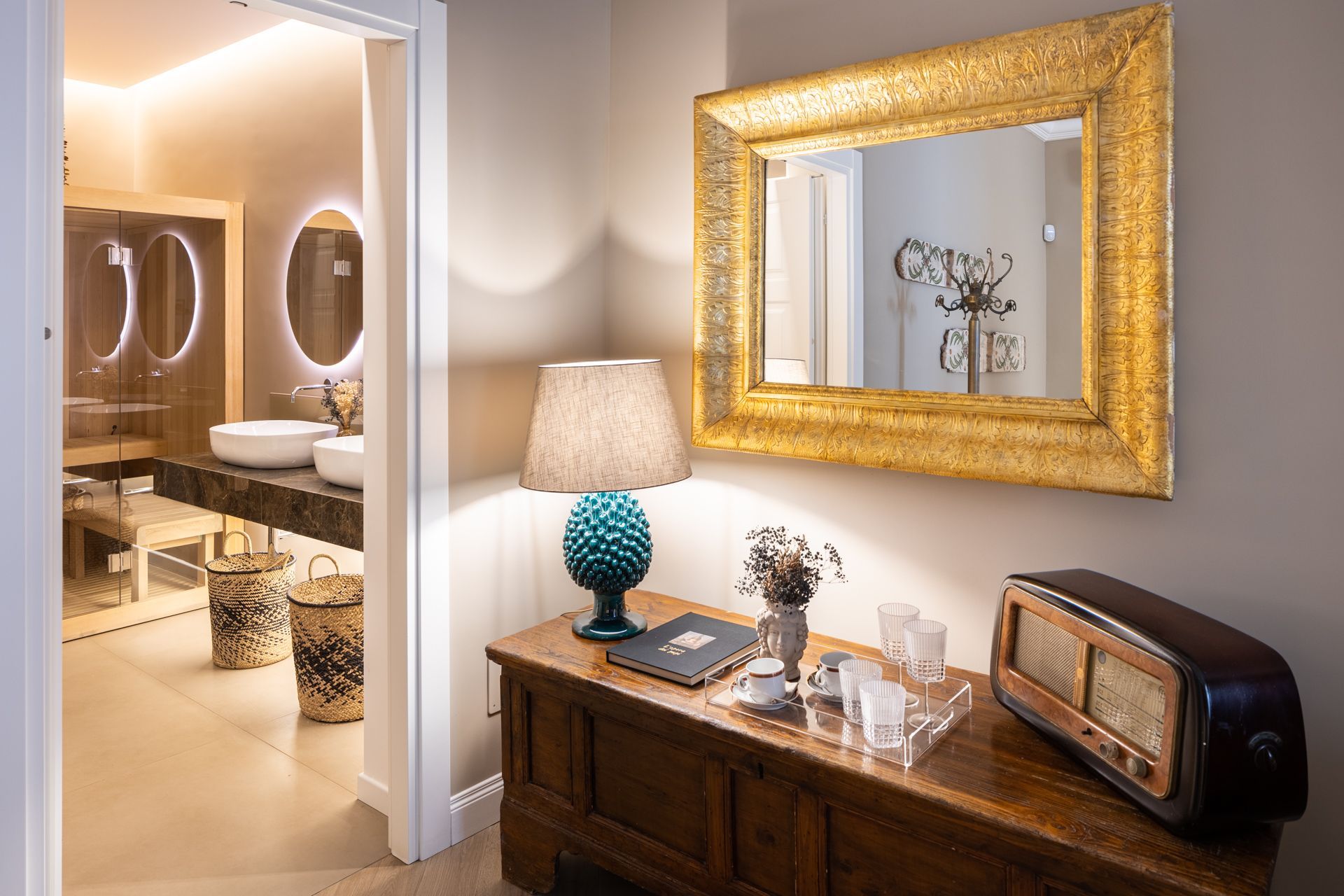 Entryway with gold-framed mirror, lamp, and vintage radio on a wooden table, leading to a modern bathroom with oval mirrors.