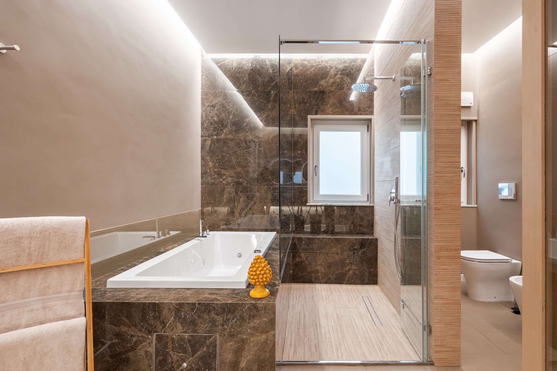 Modern bathroom with bathtub, shower, toilet, and beige/brown tile. Soft lighting.