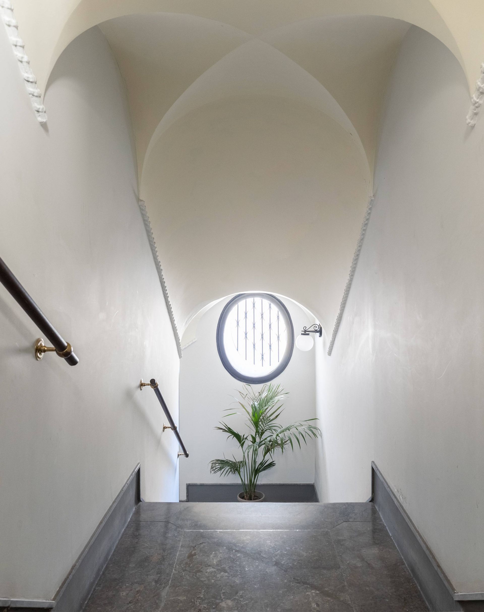 Tromba delle scale con soffitto a volta, finestra rotonda e una pianta. Pareti bianche, pavimento grigio.
