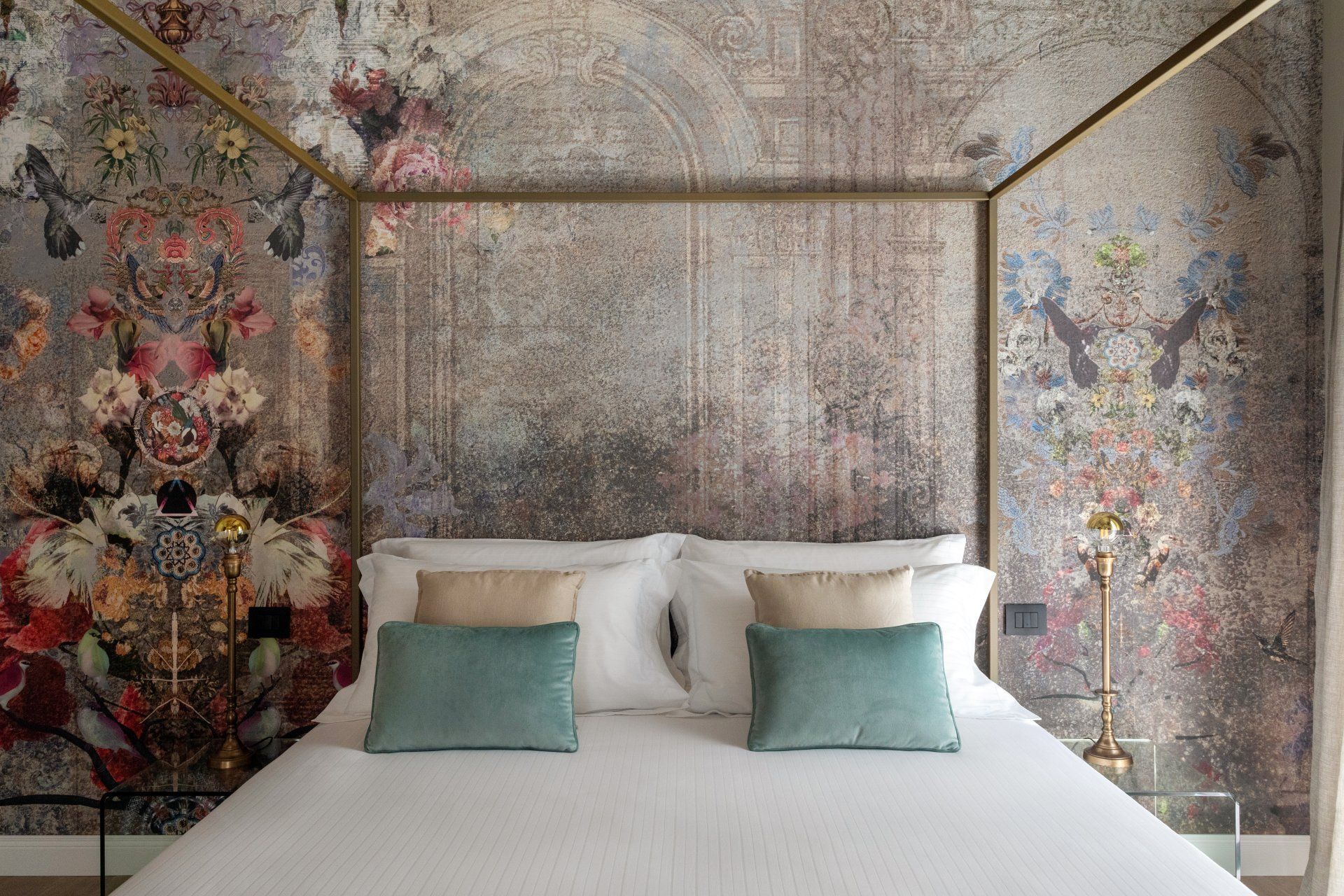 Chambre avec papier peint fleuri et lit à baldaquin. Coussins bleu sarcelle et beige sur draps blancs.