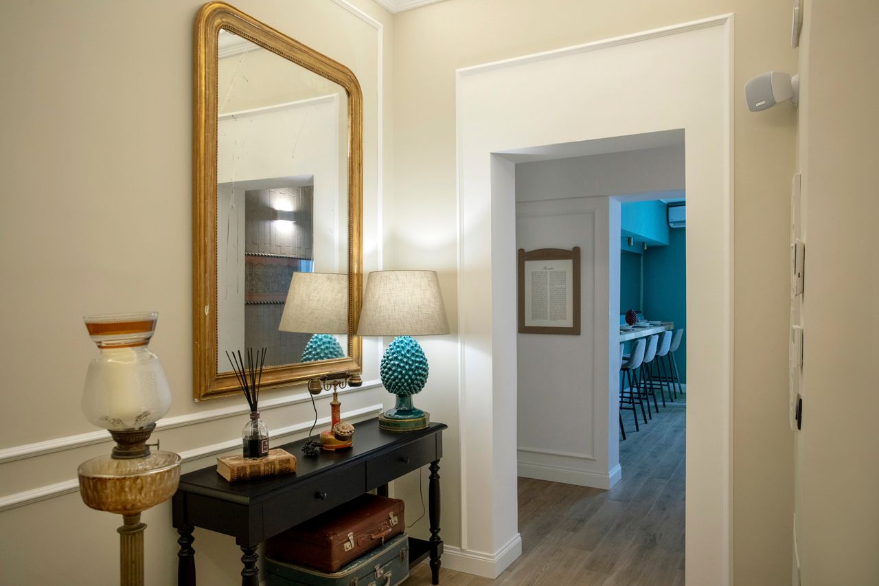 Couloir avec un miroir à cadre doré, une table noire, des lampes et une vue sur une pièce de couleur turquoise.