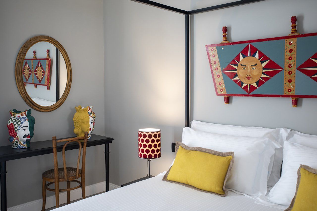 Camera da letto con letto bianco, cuscini gialli, lampada con motivi rossi, specchio e quadri colorati alle pareti.