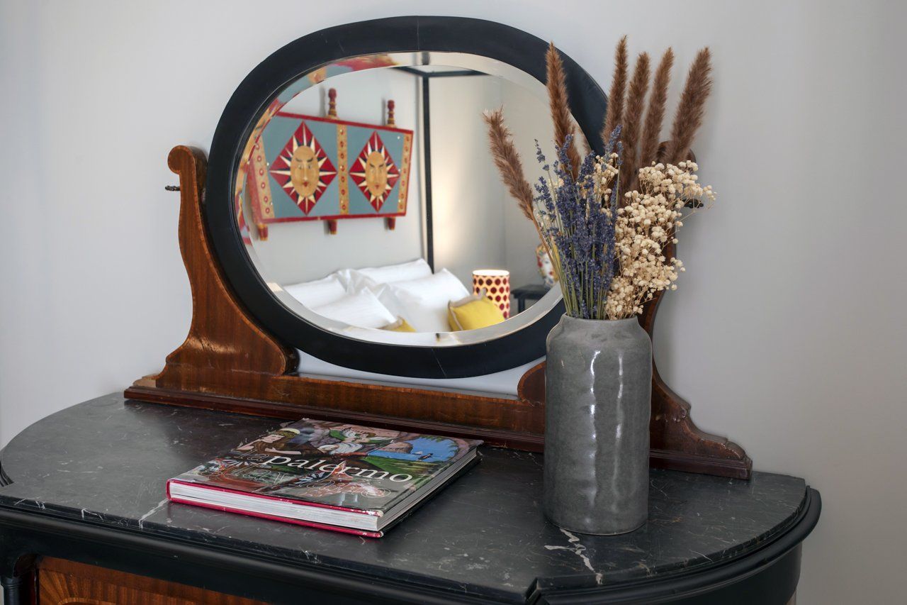 Toeletta con specchio ovale, vaso con fiori secchi e libro su un piano in marmo; riflesso del letto con decorazioni murali.