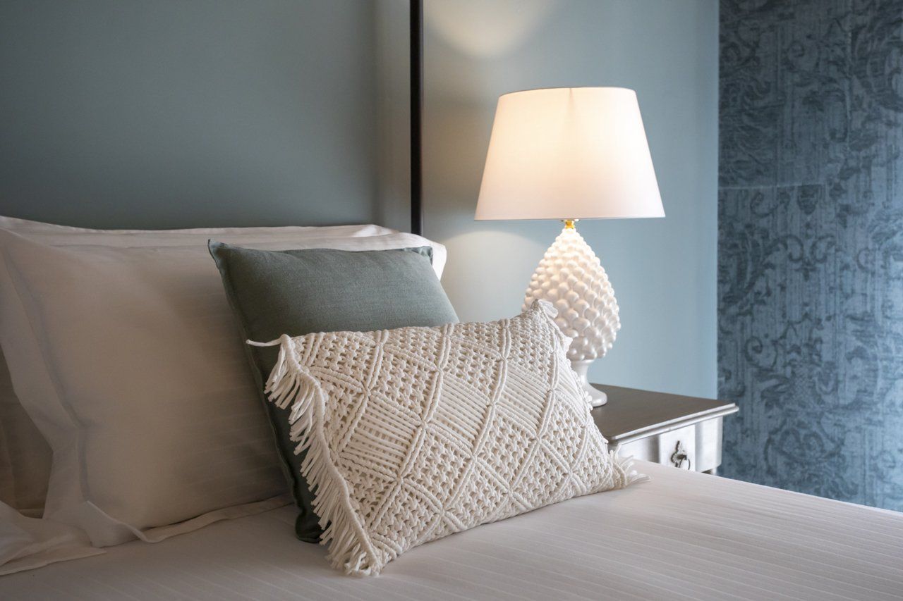 Letto con lenzuola bianche, cuscini e un cuscino decorativo in macramè, lampada da comodino, parete blu.