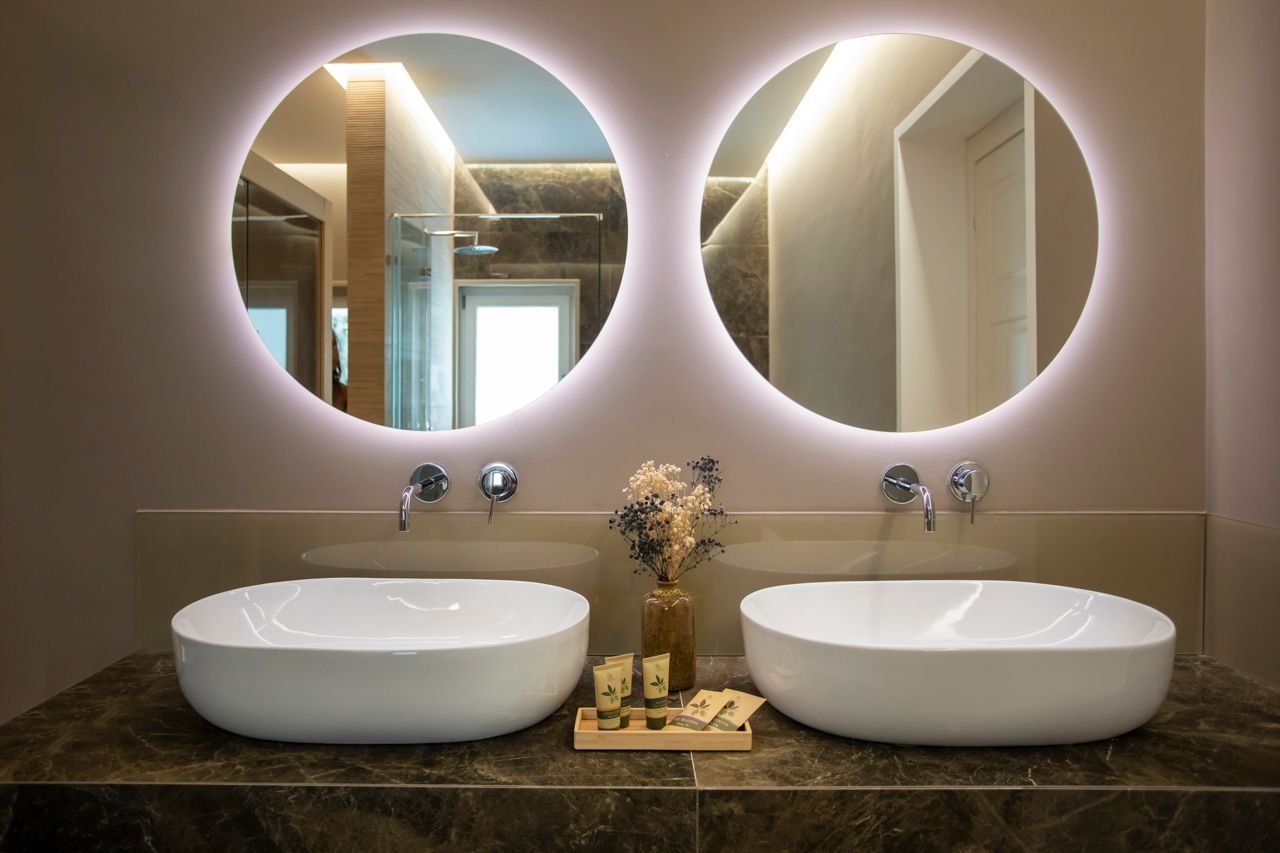 Bathroom with two round lighted mirrors, white sinks, brown countertop, and floral arrangement.