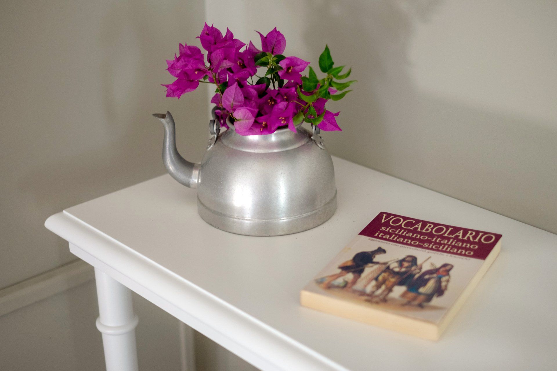 Tavolo bianco con un bollitore d'argento con fiori rosa; accanto c'è un libro con la copertina marrone.