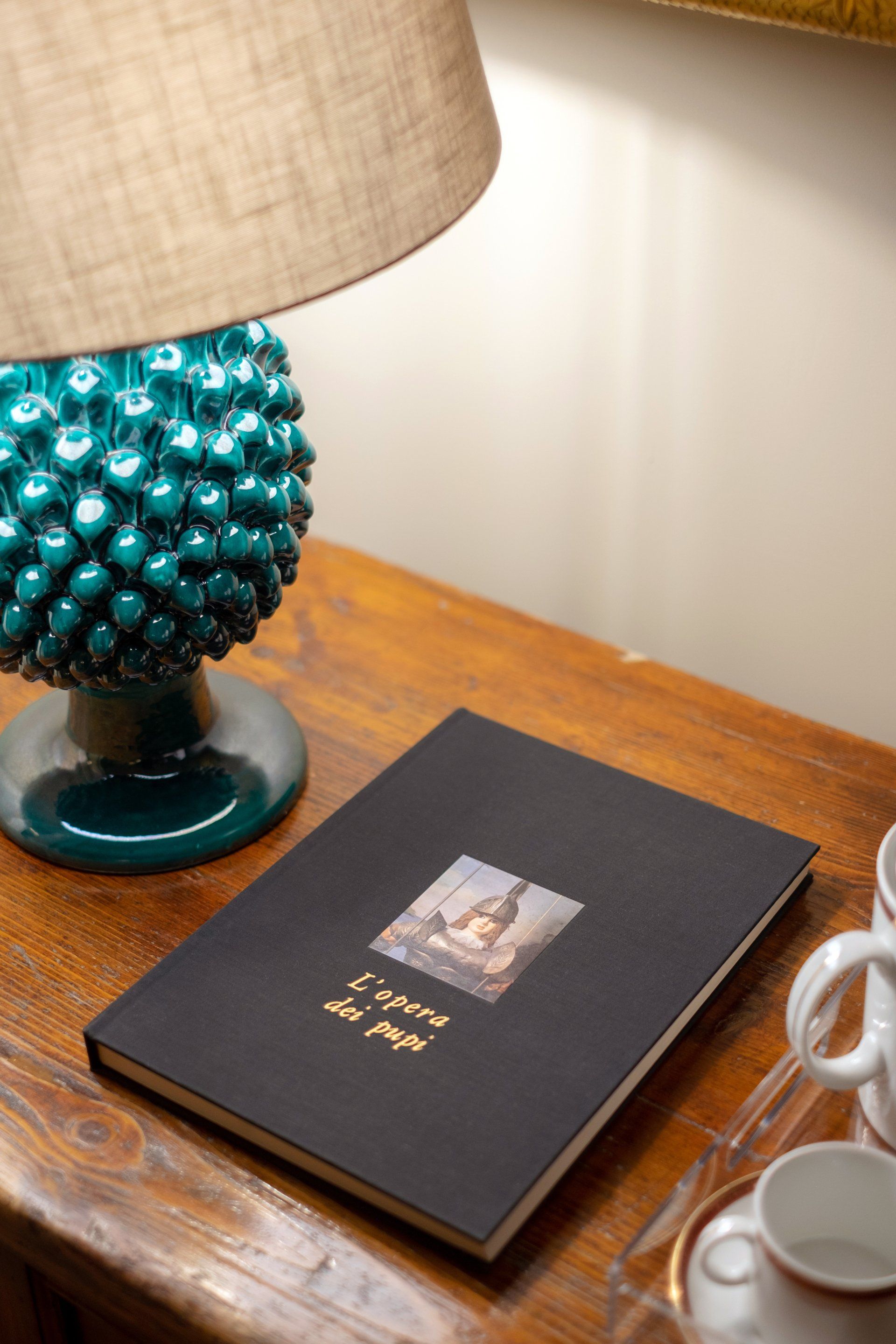 Black book on wooden table, under teal lamp with tan shade. Book has a small, central picture and gold text.