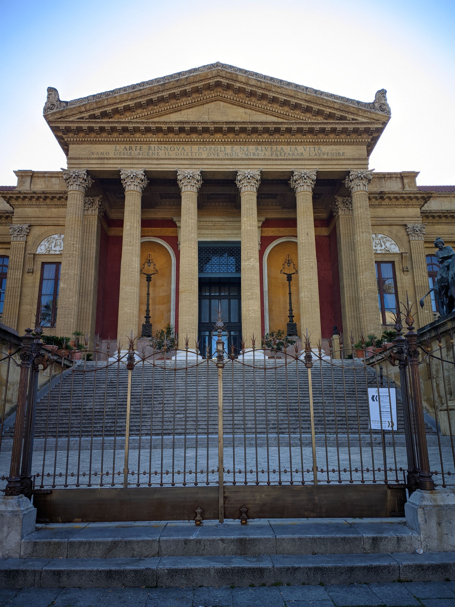 Un grand bâtiment classique beige avec des colonnes, des marches et une clôture en fer forgé noir.