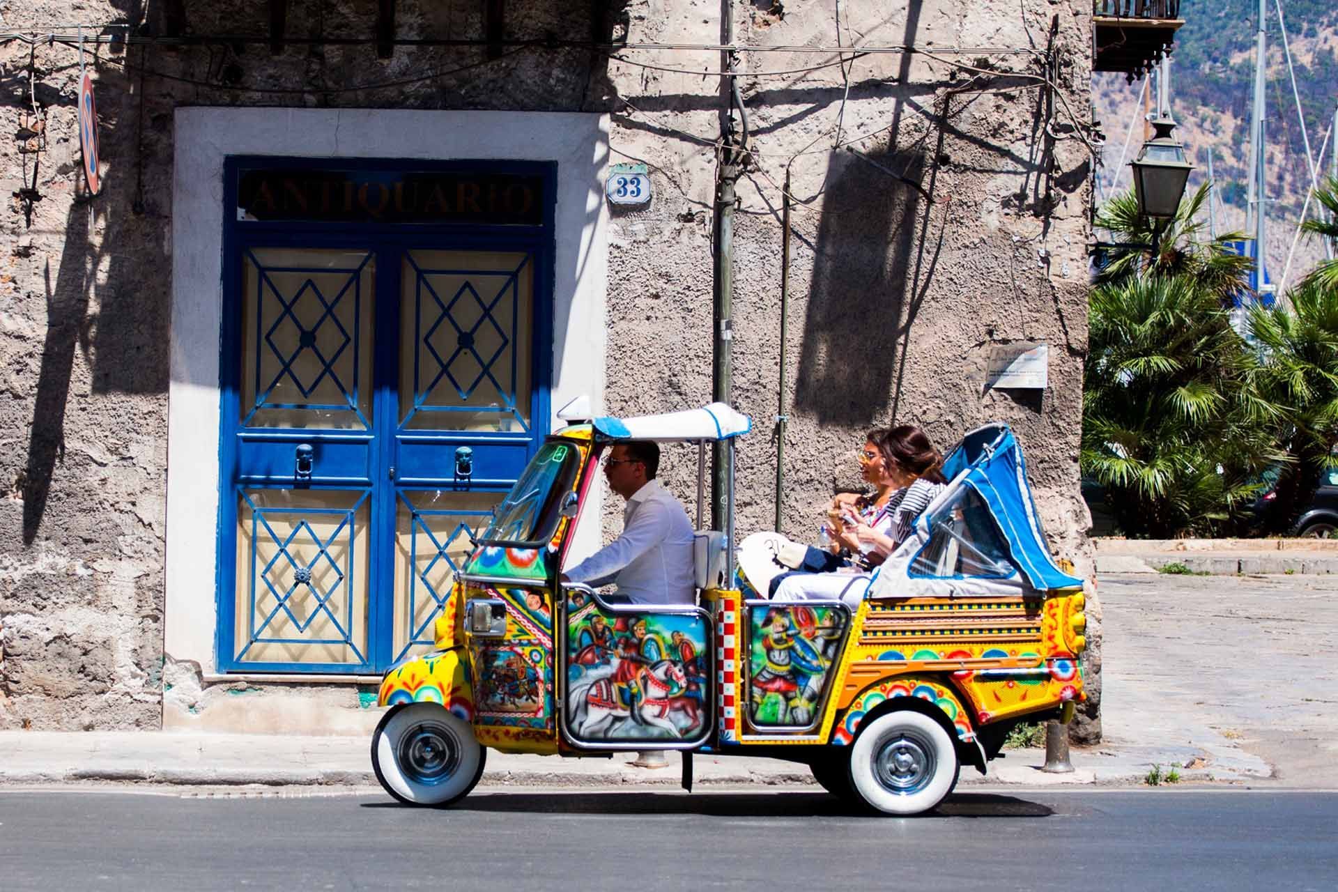 Un tuk-tuk décoré de couleurs vives transporte deux passagers et son conducteur dans une rue ensoleillée.