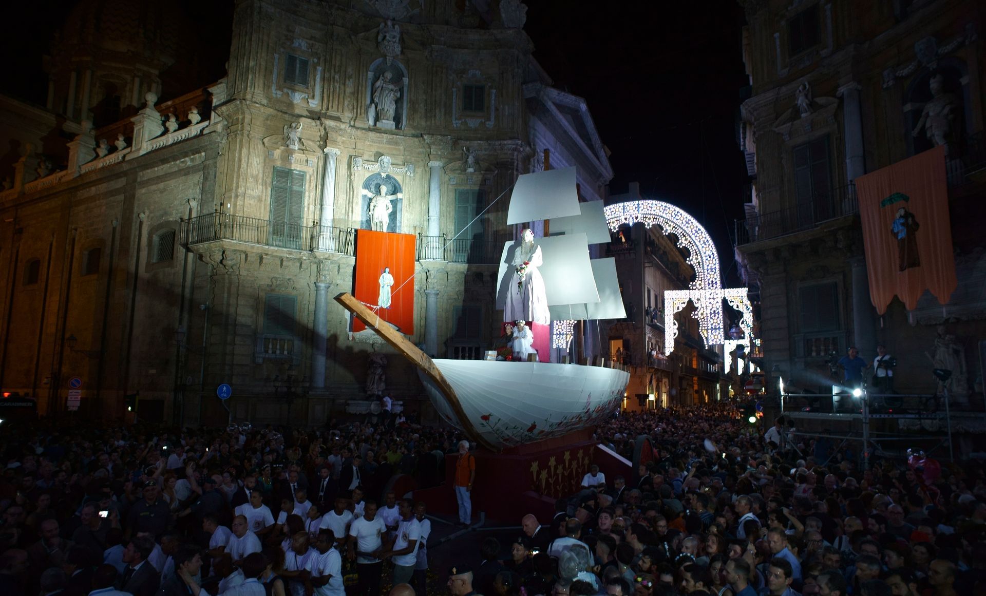 Un bateau dans une rue la nuit, une foule assiste à un spectacle avec une église en arrière-plan et des lumières vives.