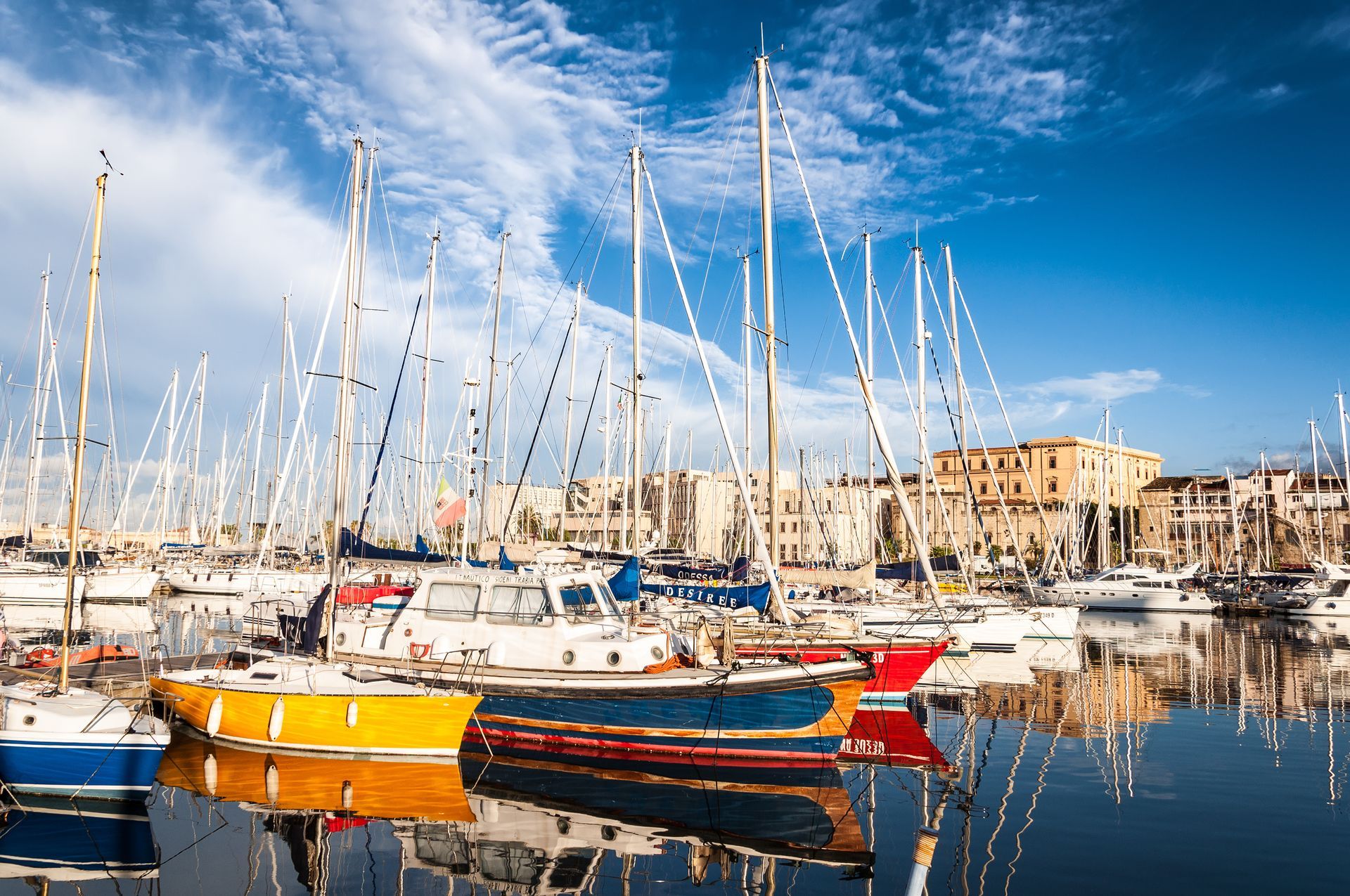 Voiliers amarrés dans un port, couleurs éclatantes, ciel bleu et bâtiments en arrière-plan.