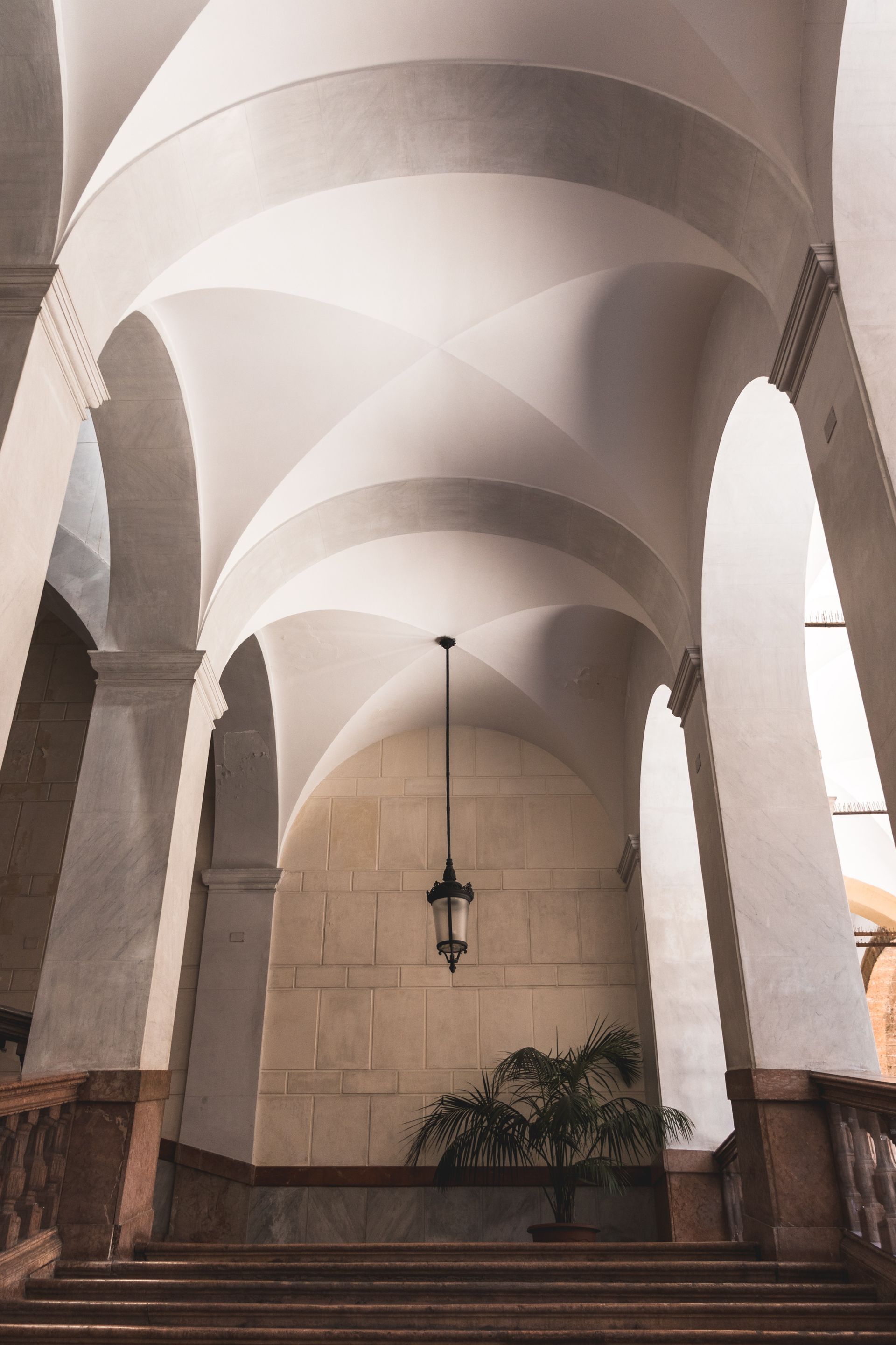 Couloir voûté avec colonnes en pierre, marches et luminaire suspendu. Une plante est posée au fond.