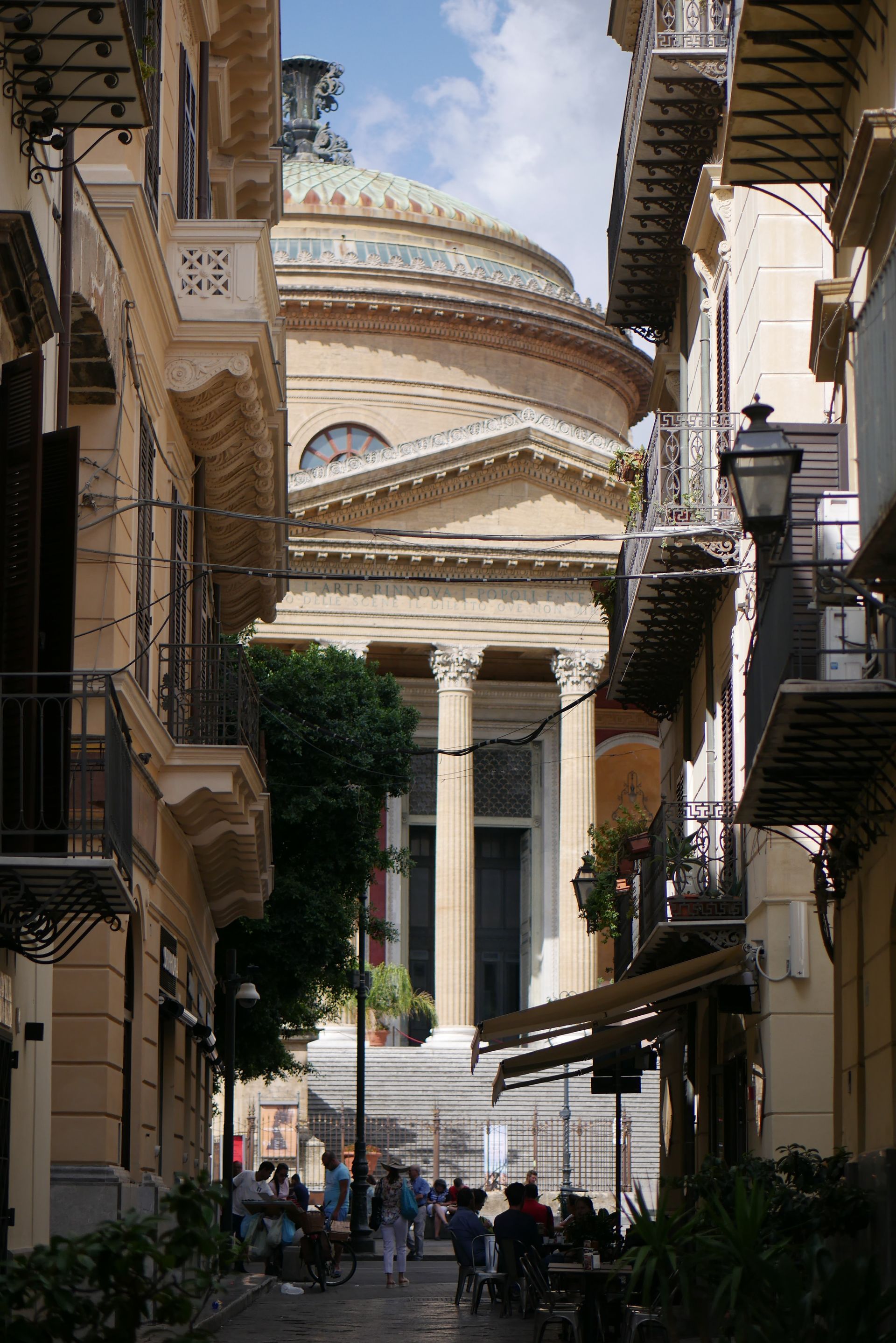 Vue d'une rue étroite donnant sur un bâtiment classique surmonté d'un dôme ; des gens marchent dans la rue et sont assis à des tables en terrasse.