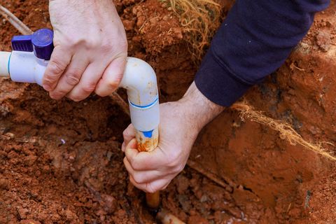 Hands connecting white PVC pipes in a dirt trench during plumbing work