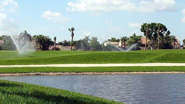 Lush green golf course with sprinklers spraying near a water hazard under a blue sky