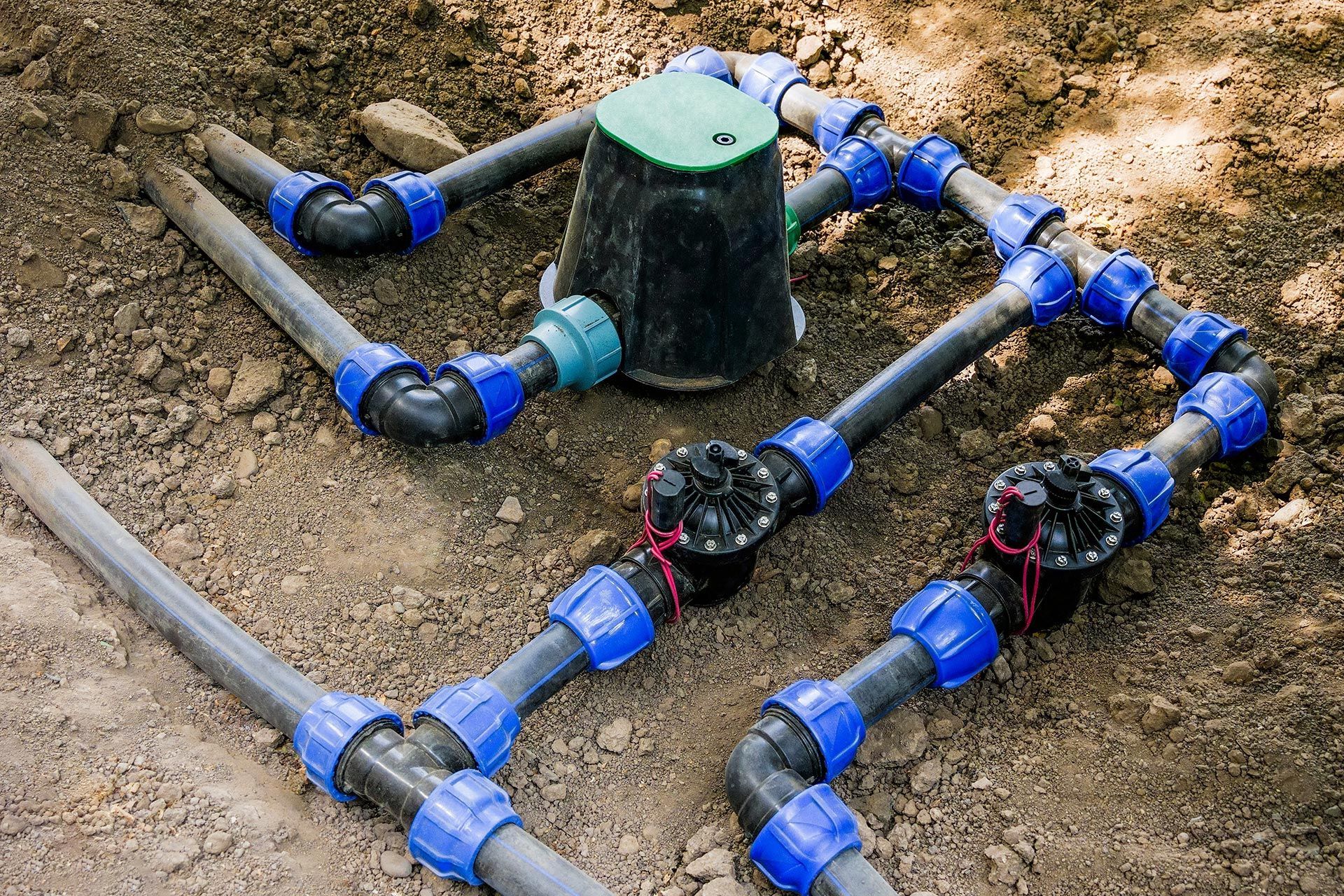 Plastic irrigation pipes and blue valves connected around a green sprinkler head in a dirt field
