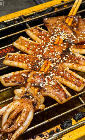Grilled squid skewered on a barbecue, coated with sauce and sesame seeds.