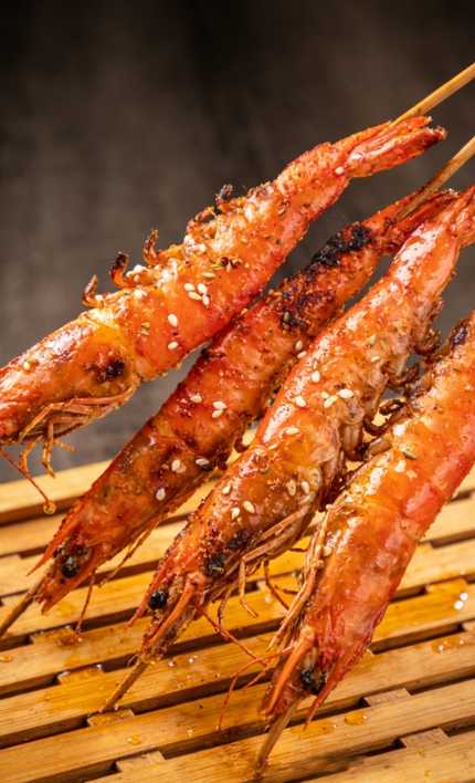 Grilled prawns skewered over a bamboo mat, browned and glistening with sauce.