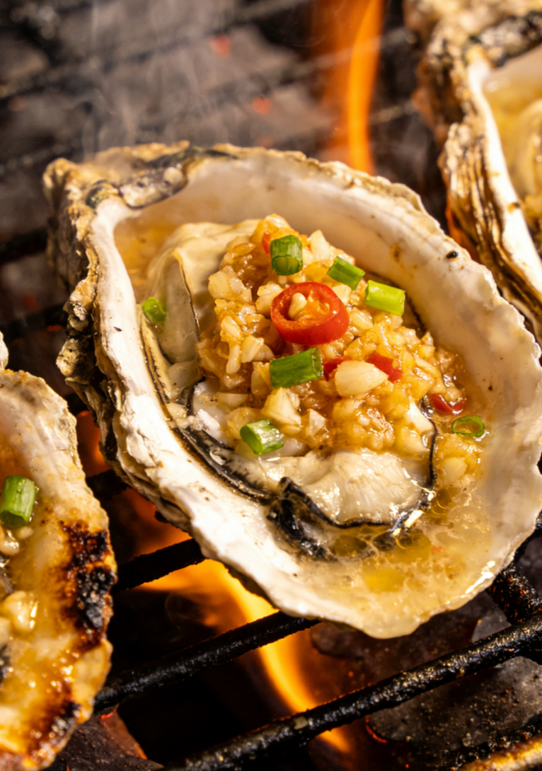 Grilled oysters topped with garlic, green onions, and red chili on an open flame