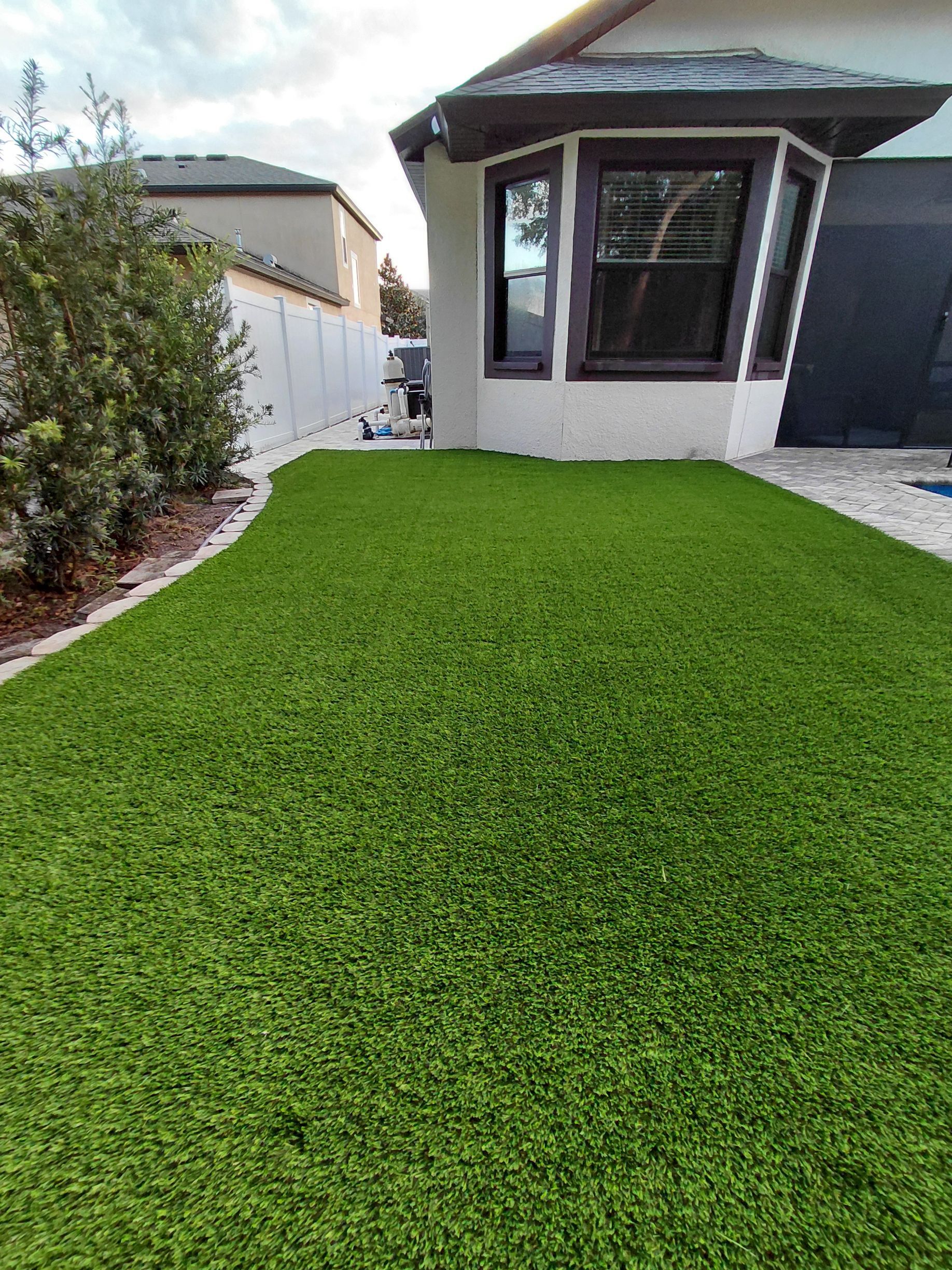 Backyard with bright green artificial turf leading to a house with a bay window and side fence