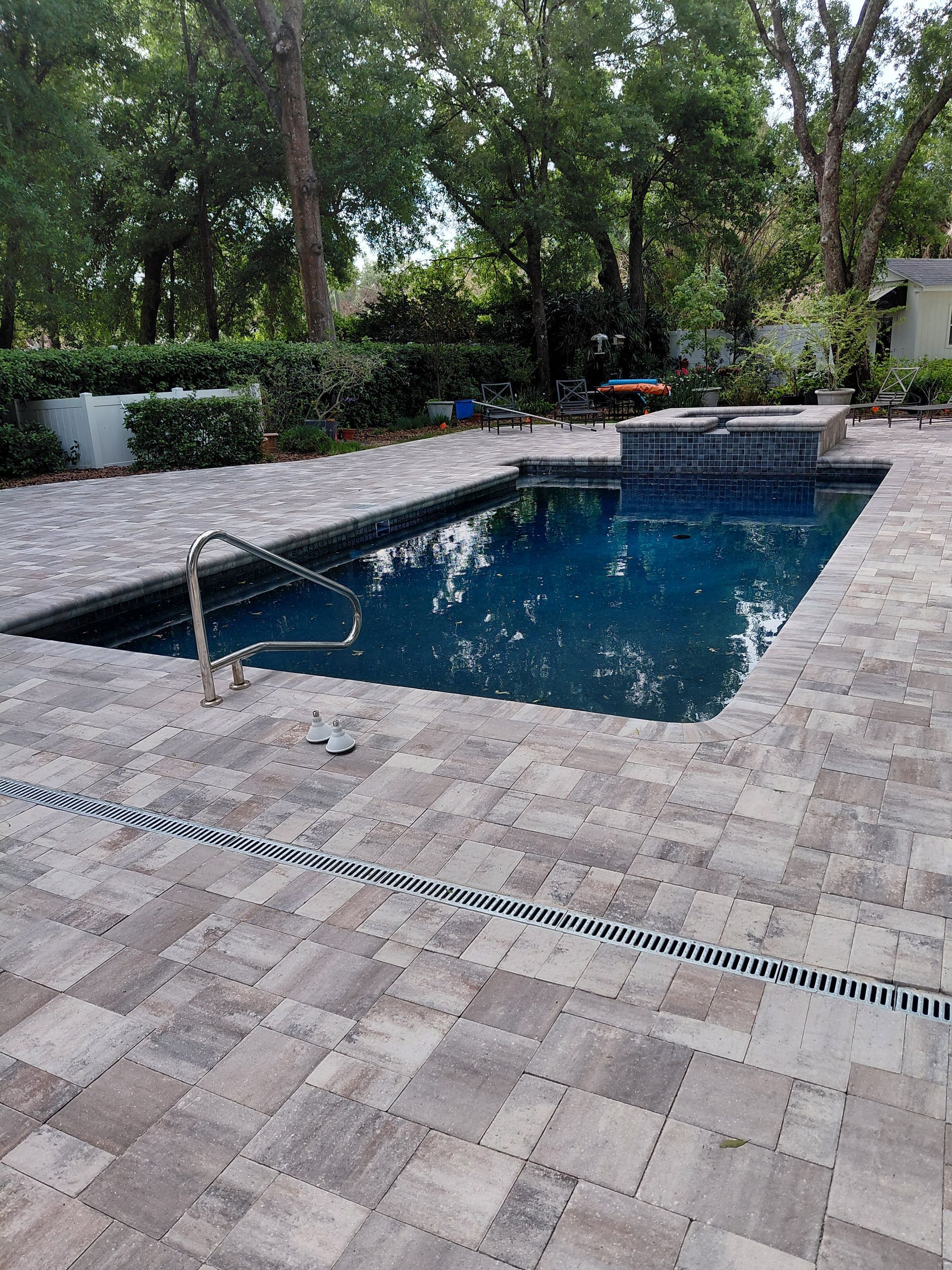 Stone-paver patio with a rectangular pool and surrounding trees, viewed from the deck