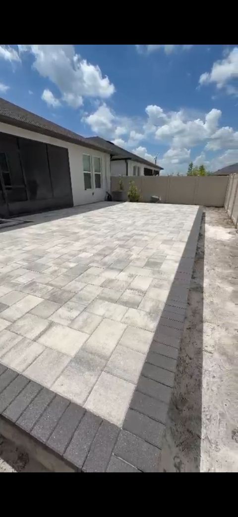 Backyard patio with light gray pavers beside a house and white wall under a blue sky with clouds