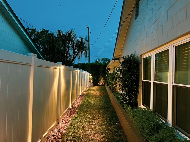 A narrow, grassy pathway between a white vinyl fence illuminated by wall lights and the side of a house at twilight. A narrow, grassy pathway between a white vinyl fence illuminated by wall lights and the side of a house at twilight.