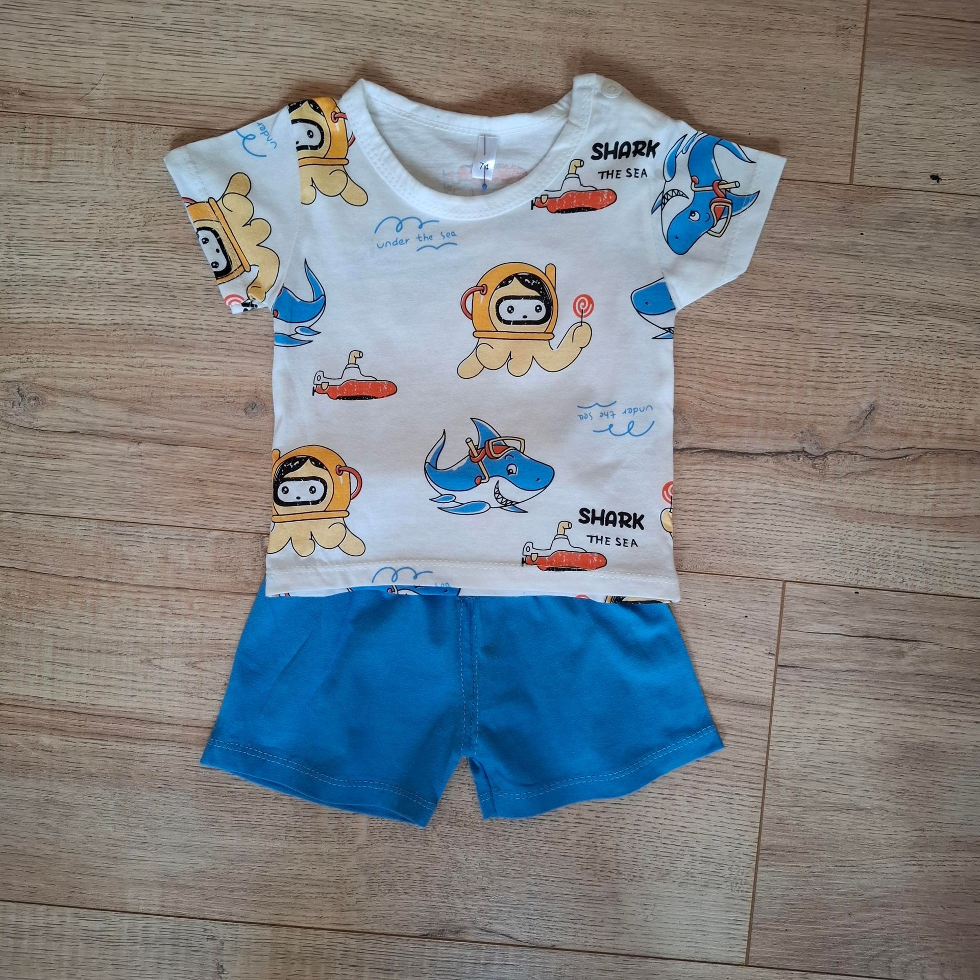 Witte en blauwe kinderpyjama met cartoonafbeeldingen van een leeuw en een haai, platliggend op een houten vloer.