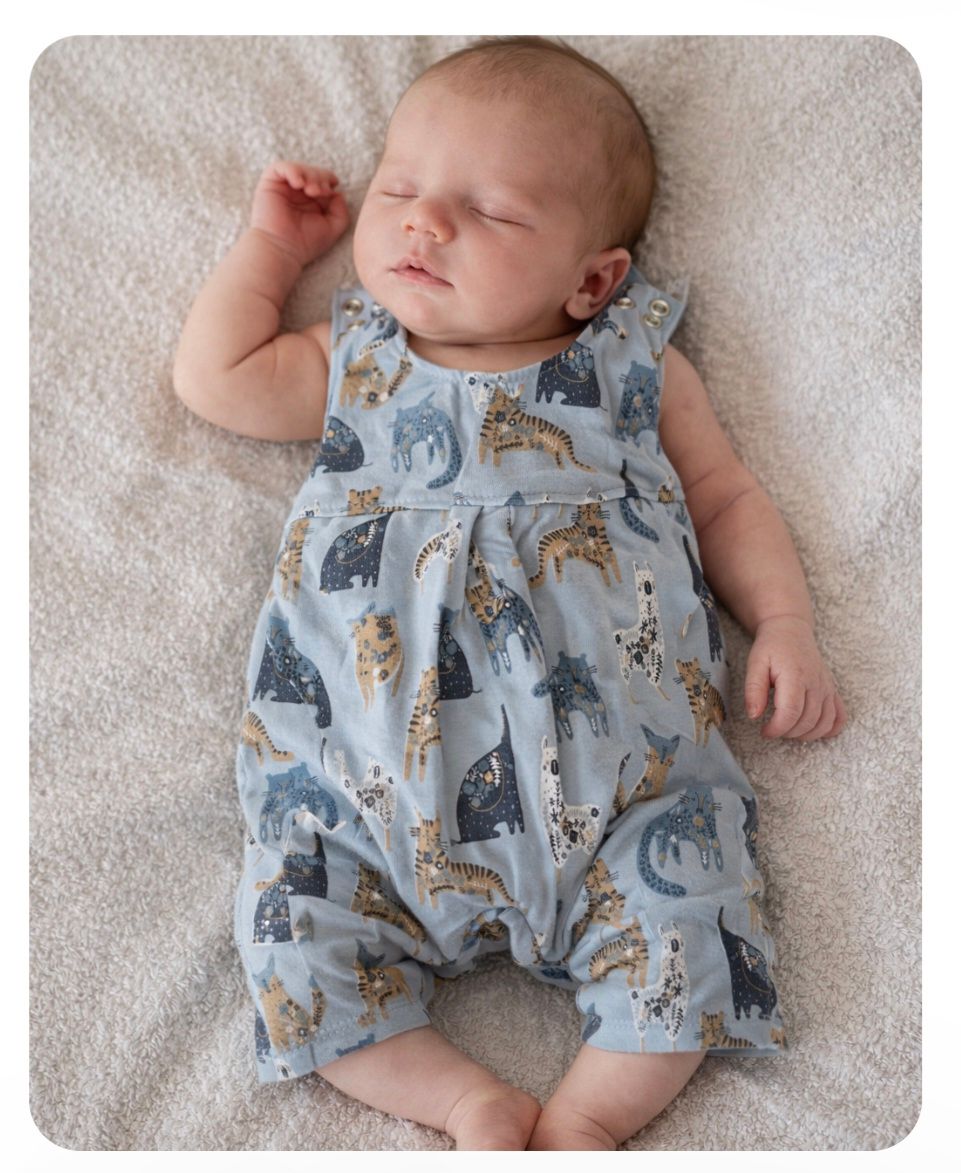 Een slapende baby in een lichtblauw rompertje met patroon ligt op een zachte witte deken met de armen omhoog.