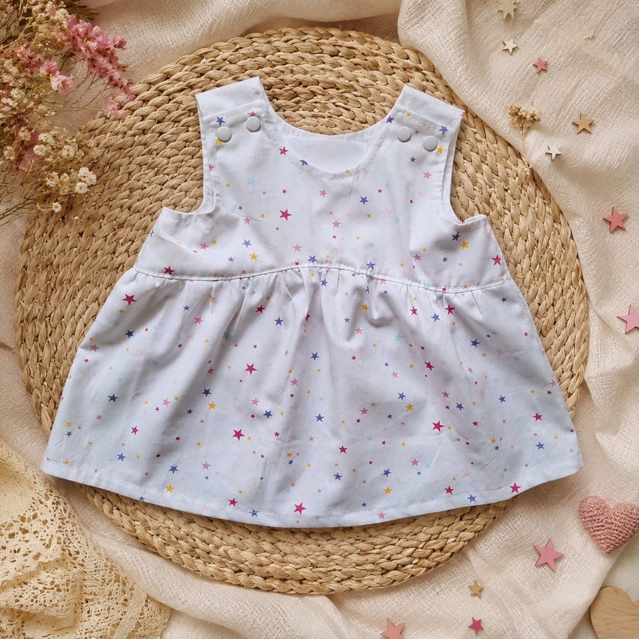 Witte mouwloze babyjurk met kleine pastelkleurige sterretjes op een geweven mat.