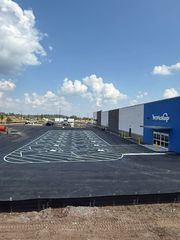 Outdoor construction lot with black surface, white markings, and a blue building under a clear sky