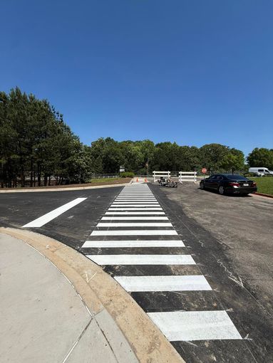 Crosswalk leading across a gravel lot beside trees, with parked cars and a clear blue sky.