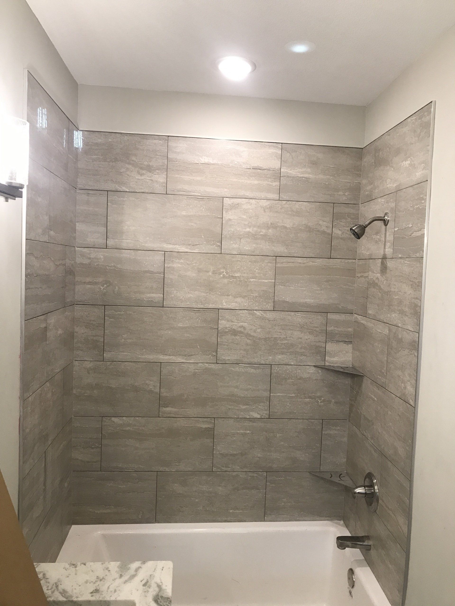 Shower with gray tile walls, fixtures installed, and a white bathtub.